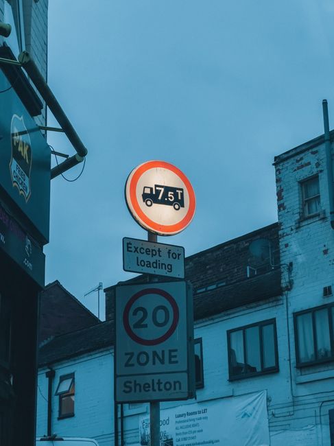 A vertical street signpost featuring three signs against a background of urban buildings and a cloudy sky. The top sign indicates a weight restriction of 7.5 tonnes for vehicles, with an exception for loading purposes listed on a smaller rectangular sign below. The middle circular sign displays a speed limit of 20 miles per hour for a designated zone, with a label specifying the zone as 'Shelton.' The street scene is typical of an urban environment, and the signage is relevant to vehicle access considerations for house removal and furniture transport services provided by Man and Van Kennington, focusing on efficient relocation logistics within residential areas.