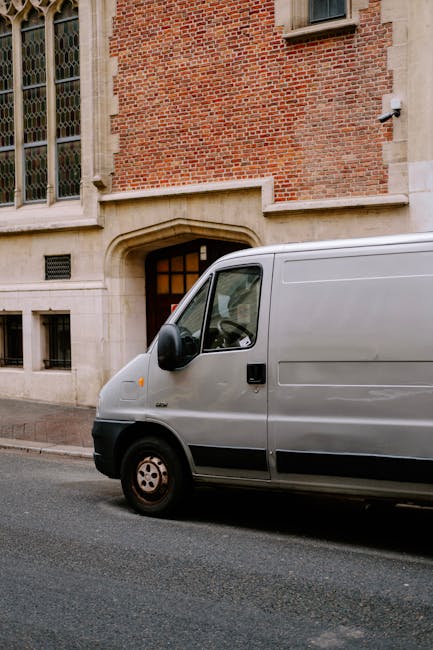 A grey delivery van parked on a paved street in front of a historic red brick building with large stone window sills and decorative stonework around the arched entrance. The van's side door and front window are visible, with the vehicle positioned close to the building's entrance. The scene is captured during daytime with natural lighting, and the surrounding environment suggests a residential or historic area suitable for home relocation and furniture transport. Occasionally, Man and Van Kennington offers removals and packing services that involve such loading and unloading processes, often utilizing vans similar to the one pictured for efficient house removals.