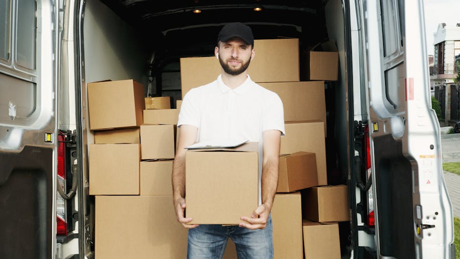 A man with a beard, wearing a black cap, white shirt, and jeans, is standing inside the open back of a large white van during a home relocation process. He is holding a medium-sized cardboard box and