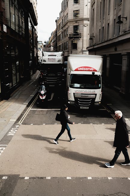 A small van with the logo and website address of Man and Van Kennington is parked on a narrow urban street, positioned behind a larger white delivery truck marked with 'Becton Transport.' The scene shows a loading area with a white motorbike parked nearby and pavement markings indicating a designated loading zone. Two pedestrians, one in dark clothing and the other in a dark suit, are crossing the street, casting distinct shadows on the pavement under an overcast sky. The street is lined with multi-storey buildings with large windows and fire escapes, typical of a city environment. The scene captures a moment during house or furniture removals, with items possibly being loaded or unloaded, and reflects a typical logistics operation with vehicles oriented for home relocation or furniture transport, supported by the presence of moving-related equipment and urban surroundings. Man and Van Kennington's services are implied through the vehicle signage, fitting with the context of packing and moving activities.