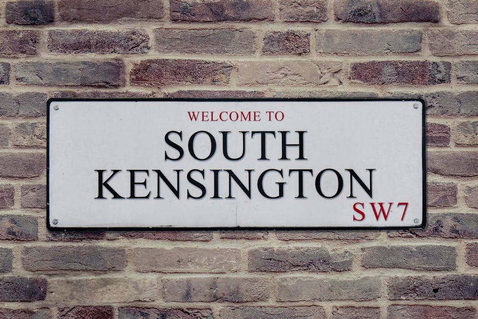 A rectangular white metal street sign mounted on a red brick wall, displaying the text 'Welcome to South Kensington SW7' in black and red lettering. The sign is positioned at about eye level and is secured with four screws, one in each corner. The brick wall has a rough texture with visible mortar lines between the bricks, which vary in shades of red and brown. The background suggests an exterior residential or commercial building in South Kensington. This setting may depict the entrance or a facade where a house removal or relocation service might operate, relevant to the context of professional moving and packing services provided by Man and Van Kennington involved in home relocations and furniture transport.