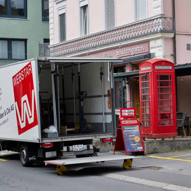 A small van with the logo and website address of Man and Van Kennington is parked on a narrow urban street, positioned behind a larger white delivery truck marked with 'Becton Transport.' The scene shows a loading area with a white motorbike parked nearby and pavement markings indicating a designated loading zone. Two pedestrians, one in dark clothing and the other in a dark suit, are crossing the street, casting distinct shadows on the pavement under an overcast sky. The street is lined with multi-storey buildings with large windows and fire escapes, typical of a city environment. The scene captures a moment during house or furniture removals, with items possibly being loaded or unloaded, and reflects a typical logistics operation with vehicles oriented for home relocation or furniture transport, supported by the presence of moving-related equipment and urban surroundings. Man and Van Kennington's services are implied through the vehicle signage, fitting with the context of packing and moving activities.