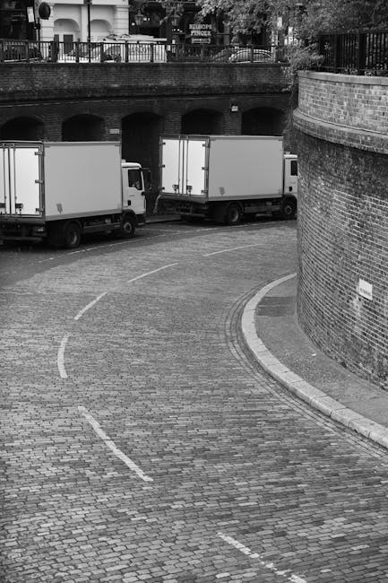 A black and white photograph of a small, curved cobblestone street in an urban setting during daytime, with three white box trucks parked along the side. The trucks are positioned parallel with their