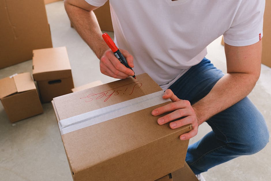 A man with dark curly hair and a beard, dressed in a navy t-shirt with a blue patterned pocket and dark trousers, is inside a bright, empty room with white walls and light wooden flooring. He is holdi