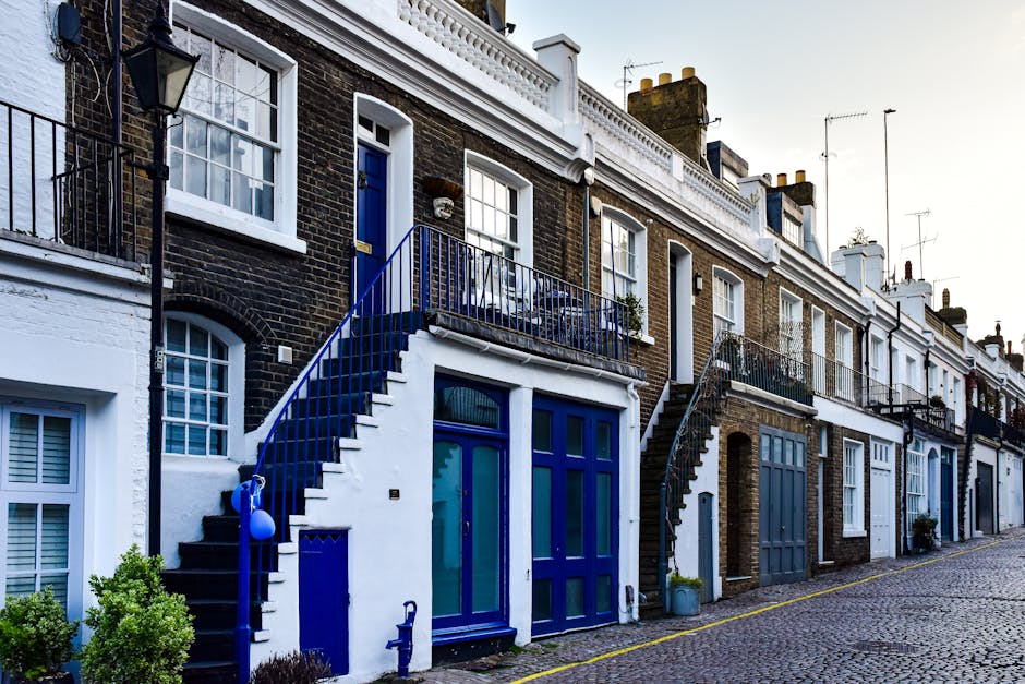 A row of terraced residential buildings with brick facades, some painted white, featuring large windows with white frames. The ground level includes blue doors and large windows, with external staircases leading to upper-floor entrances; one staircase is painted bright blue with a matching blue railing, and another has black steps with a black railing. Potted plants and small shrubs are positioned near the entrances. The street is paved with cobblestones, and there are no vehicles visible in the image. The lighting suggests daytime with a clear sky, and the scene reflects typical urban house frontage suitable for household relocation and furniture transport, with visible elements indicating ongoing home moving or packing activities.