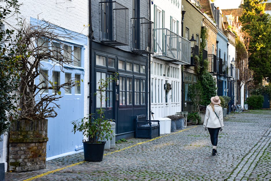 A woman in a beige coat, black pants, and a wide-brimmed hat walking along a cobbled street lined with terraced houses. The houses have mixed exterior finishes of white, blue, and dark grey, with some featuring small balconies and potted plants. Several black and white bins are placed outside, along with a black bench and various cardboard boxes used for packing during a home relocation. A large potted plant is positioned near a light blue door, and bicycles are visible parked against the buildings. The scene is illuminated by natural daylight, with trees and other greenery in the background, suggesting a residential area suitable for furniture transport, packing and moving, or loading and unloading activities related to house removals. This image captures the typical environment where Man and Van Kennington may assist with house removals and moving logistics, reflecting the process of loading items onto a vehicle outside a property.