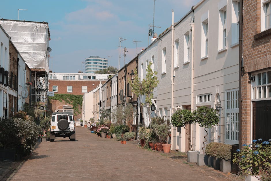 A quiet residential street lined with terraced houses on both sides, featuring white and brick exterior facades, with potted plants and small trees arranged along the pavement. A white van belonging to Man and Van Kennington is parked on the cobblestone street, positioned near an open doorway, indicating the ongoing process of home relocation and furniture transport. Visible inside the open doorway are cardboard boxes, wrapped furniture, and packing materials, suggesting a packing and moving operation. The street is illuminated by natural daylight, with some houses displaying window boxes, and in the background, there is a modern, circular office building contrasting with the traditional terrace homes. The scene captures the logistics involved in house removals, including the careful handling of household items, loading activities, and the preparation for transportation, aligning with services offered by Man and Van Kennington for removals and moving logistics.