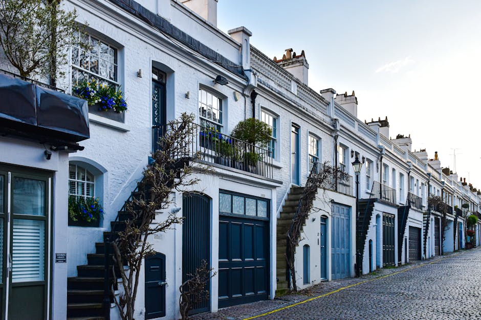 A woman in a beige coat, black pants, and a wide-brimmed hat walking along a cobbled street lined with terraced houses. The houses have mixed exterior finishes of white, blue, and dark grey, with some featuring small balconies and potted plants. Several black and white bins are placed outside, along with a black bench and various cardboard boxes used for packing during a home relocation. A large potted plant is positioned near a light blue door, and bicycles are visible parked against the buildings. The scene is illuminated by natural daylight, with trees and other greenery in the background, suggesting a residential area suitable for furniture transport, packing and moving, or loading and unloading activities related to house removals. This image captures the typical environment where Man and Van Kennington may assist with house removals and moving logistics, reflecting the process of loading items onto a vehicle outside a property.