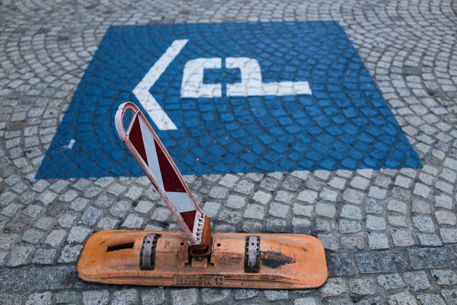 A close-up view of a cobblestone street surface showing a designated blue parking bay marked with a white arrow and a bicycle symbol, indicating it is reserved for bicycle parking. In the foreground, an orange plastic equipment base with visible signs of wear and black markings, supports a red and white striped barrier segment typically used for traffic or parking management. The barrier is slightly tilted, with the striped reflective surface facing outward. The scene is part of a home relocation or moving process, with the blue parking space and barrier indicating a temporary restriction for vehicle or equipment access. The image depicts elements relevant to house removals and logistics, where managing parking and loading zones is crucial, and is associated with professional removal services like Man and Van Kennington.