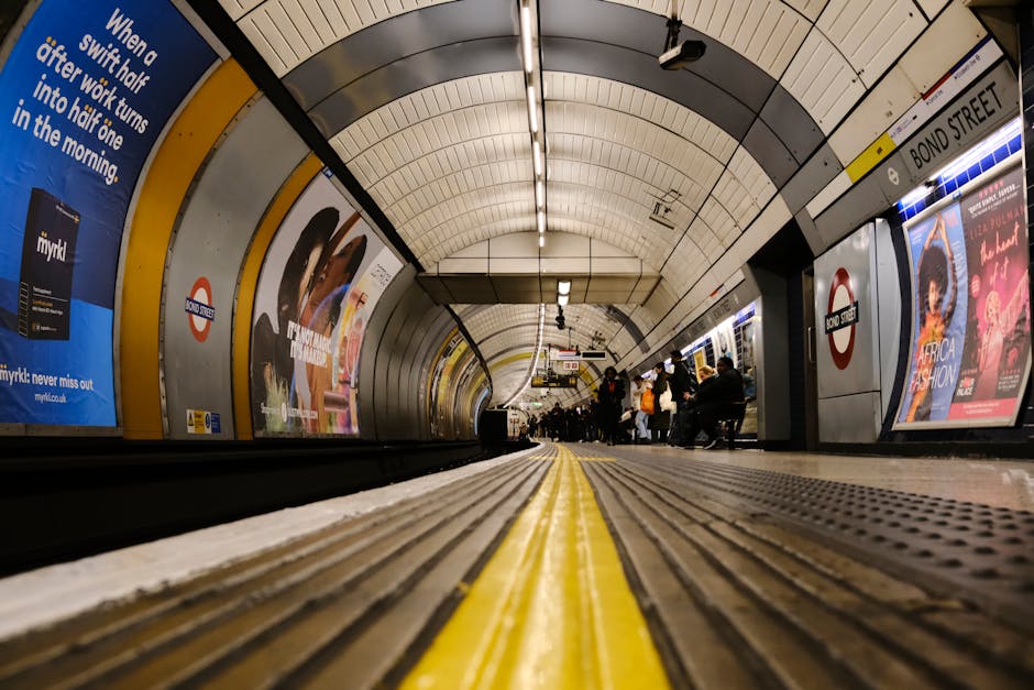A view of the London Underground platform at Oval station, taken from ground level near the edge of the platform with the yellow safety line in the foreground. The curved, tiled ceiling of the station is illuminated by fluorescent lighting, and multiple advertisements and signs are visible along the walls, including the London Underground roundel symbol above platform edge posters. Passengers are seated on benches along the platform, some holding or managing small luggage and bags, indicating preparations for home relocation or furniture transport during a moving process. The area shows a clean, structured environment typical of London Underground stations, with tiled flooring, safety markings, and a sense of ongoing activity associated with commuter travel. This setting reflects the logistical aspect of moving services provided by companies like Man and Van Kennington, involved in packing, loading, and transporting household items from urban transit hubs.