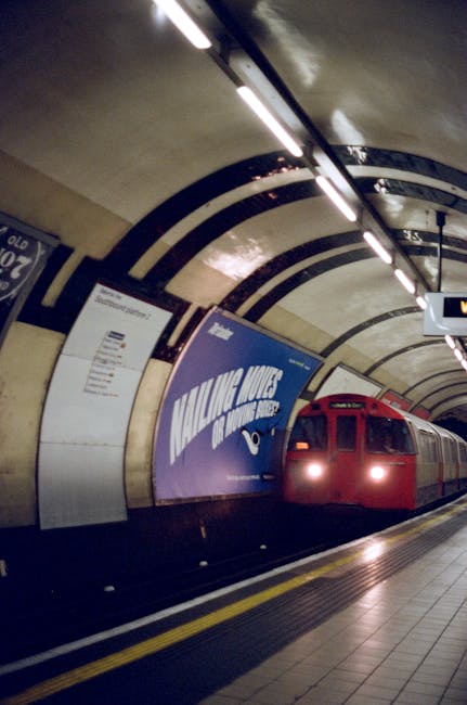 A red London Underground train is arriving at an underground station platform with a curved tunnel ceiling illuminated by fluorescent lighting. The platform has a tactile yellow safety strip along the edge, and the walls are covered with advertising posters and station information signs. In the background, the tunnel curves out of view, and a digital display board shows train departure details. The setting appears clean and well-maintained, with no visible people on the platform. This scene is related to transportation services, supporting the context of house removals and relocation logistics, such as moving services connecting commuters in Kennington, as indicated on the webpage. Man and Van Kennington often coordinates transport and packing for home relocations involving efficient furniture transport through such underground stations.