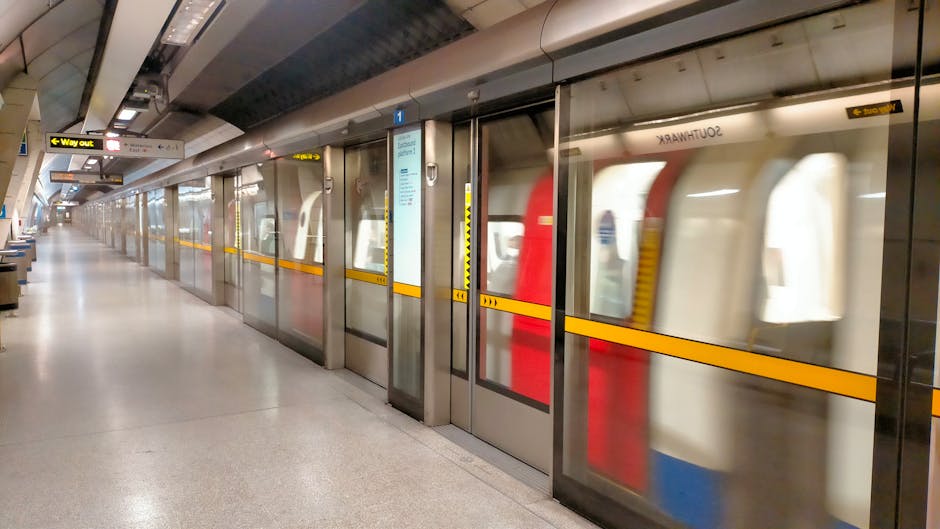 A view of the London Underground platform at Oval station, taken from ground level near the edge of the platform with the yellow safety line in the foreground. The curved, tiled ceiling of the station is illuminated by fluorescent lighting, and multiple advertisements and signs are visible along the walls, including the London Underground roundel symbol above platform edge posters. Passengers are seated on benches along the platform, some holding or managing small luggage and bags, indicating preparations for home relocation or furniture transport during a moving process. The area shows a clean, structured environment typical of London Underground stations, with tiled flooring, safety markings, and a sense of ongoing activity associated with commuter travel. This setting reflects the logistical aspect of moving services provided by companies like Man and Van Kennington, involved in packing, loading, and transporting household items from urban transit hubs.