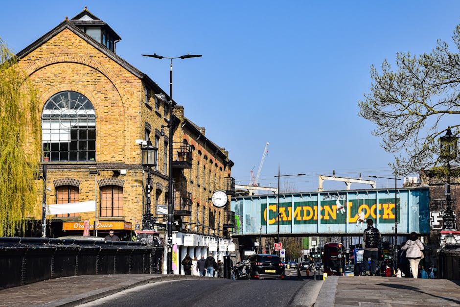 A street scene in Kennington featuring a yellow brick building with large arched windows and a black roof. In the foreground, pedestrians are walking along the pavement, some carrying bags. A black van is parked nearby, and a blue and yellow Caidin Lock freight train bridge crosses the street. Streetlights and a clock are mounted on black poles along the sidewalk. To the right, a leafless tree with sparse branches is visible against a clear blue sky. The environment suggests an active urban area during daylight hours, with the presence of vehicles, pedestrians, and construction cranes in the background indicating ongoing development. This scene is related to house removals or moving logistics, as it captures typical urban features involved in furniture transport and packing and moving activities, relevant to services like those offered by Man and Van Kennington.