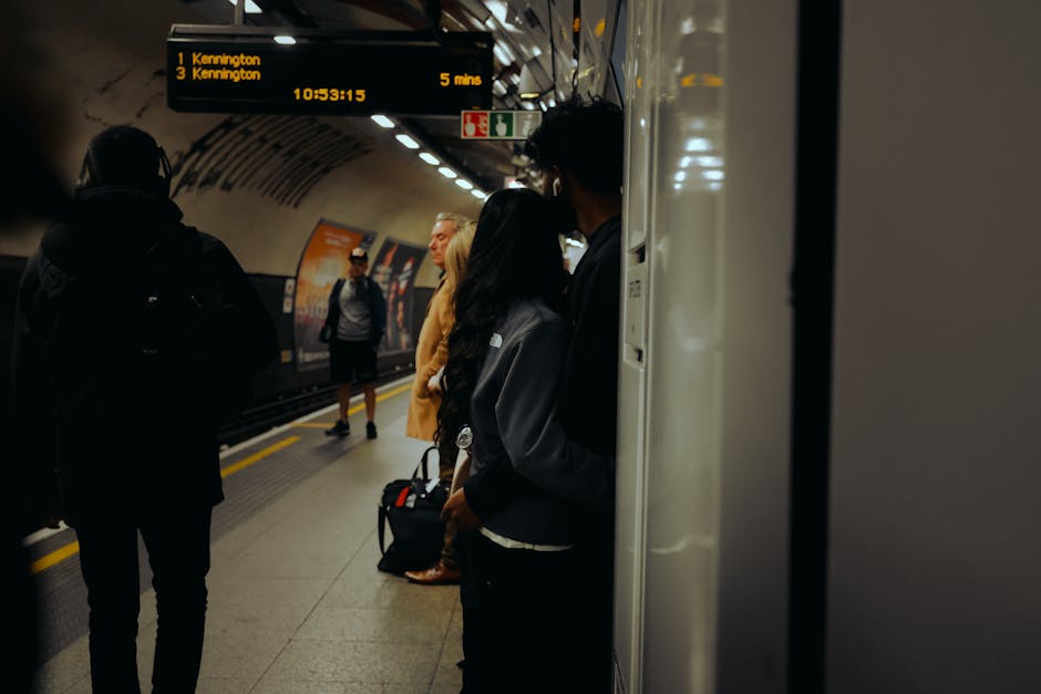 Inside an underground train station, several passengers are waiting for their train. Some are standing near the platform edge, with a black backpack and a small carry-on visible on the ground. One person is positioned next to the train door, partially obscured by the door frame, and appears to be holding a bag or suitcase. Opposite, a man wearing a dark jacket and glasses stands facing away from the camera, with other commuters visible further along the platform. An electronic display board overhead shows scheduled train arrivals for Kennington, indicating delays and platform information, with the time displayed as 10:53. The station environment features modern lighting, tiled walls, and a curved ceiling. This scene reflects the typical process of preparing for transportation, akin to home relocation logistics, with passengers waiting to board, which can be related to the planning and coordination involved in full-house removals and furniture transport services provided by Man and Van Kennington.