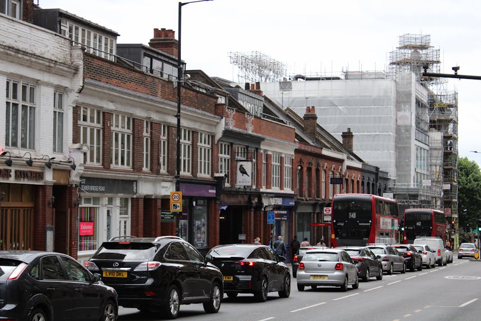 A street scene in Kennington featuring a yellow brick building with large arched windows and a black roof. In the foreground, pedestrians are walking along the pavement, some carrying bags. A black van is parked nearby, and a blue and yellow Caidin Lock freight train bridge crosses the street. Streetlights and a clock are mounted on black poles along the sidewalk. To the right, a leafless tree with sparse branches is visible against a clear blue sky. The environment suggests an active urban area during daylight hours, with the presence of vehicles, pedestrians, and construction cranes in the background indicating ongoing development. This scene is related to house removals or moving logistics, as it captures typical urban features involved in furniture transport and packing and moving activities, relevant to services like those offered by Man and Van Kennington.