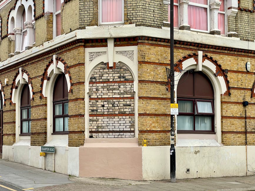 Photograph of the exterior corner of a Victorian-style brick building on a London street, featuring arched windows with dark brown frames and pink curtains, and decorative white architectural detailing above the windows and along the façade. The ground level has a white-painted base with a small, pinkish-painted step leading to a doorway that is not visible. A black lamppost with a yellow and white street sign attached is positioned at the corner, and a metal pole with signage is also present near the curb. The pavement is clean, and the scene suggests a typical urban setting suitable for house removals or furniture transport, with no visible interior or moving activity present.