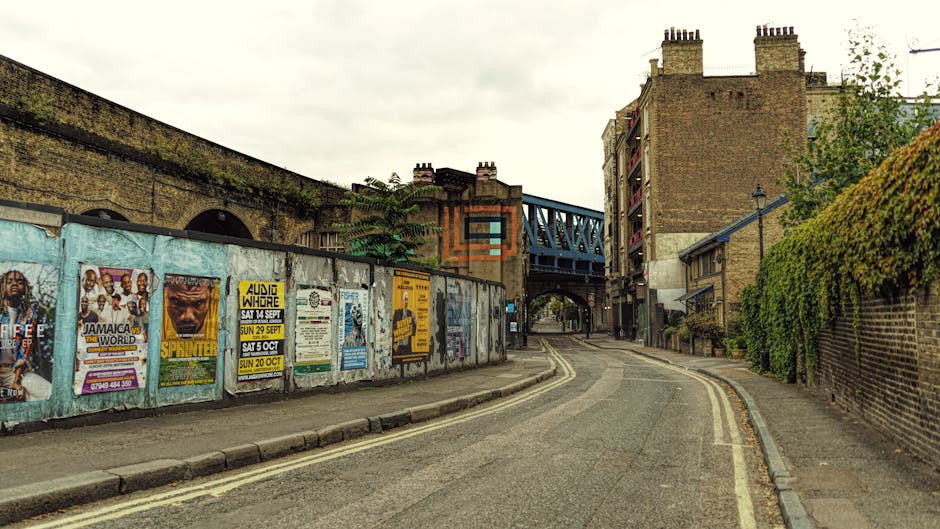 A view of a row of traditional three-storey terraced houses with brick facades, white-framed sash windows, and black iron railings along the pavement, situated on Kennington Lane in London. The buildings have a uniform appearance with decorative stone lintels above the windows and slate roofs with dormer windows. A narrow sidewalk runs parallel to the street, which appears to be a residential area. In front of the houses, several cardboard boxes, wrapped furniture, and packing materials are placed on the pavement, possibly prepared for a home relocation. A small moving trolley and protective blankets are visible near the entrance of one property, indicating ongoing packing or loading activities. Overall, the scene depicts a typical London residential street during a house move, with elements of furniture transport and packing materials visible as part of the moving process carried out by Man and Van Kennington.